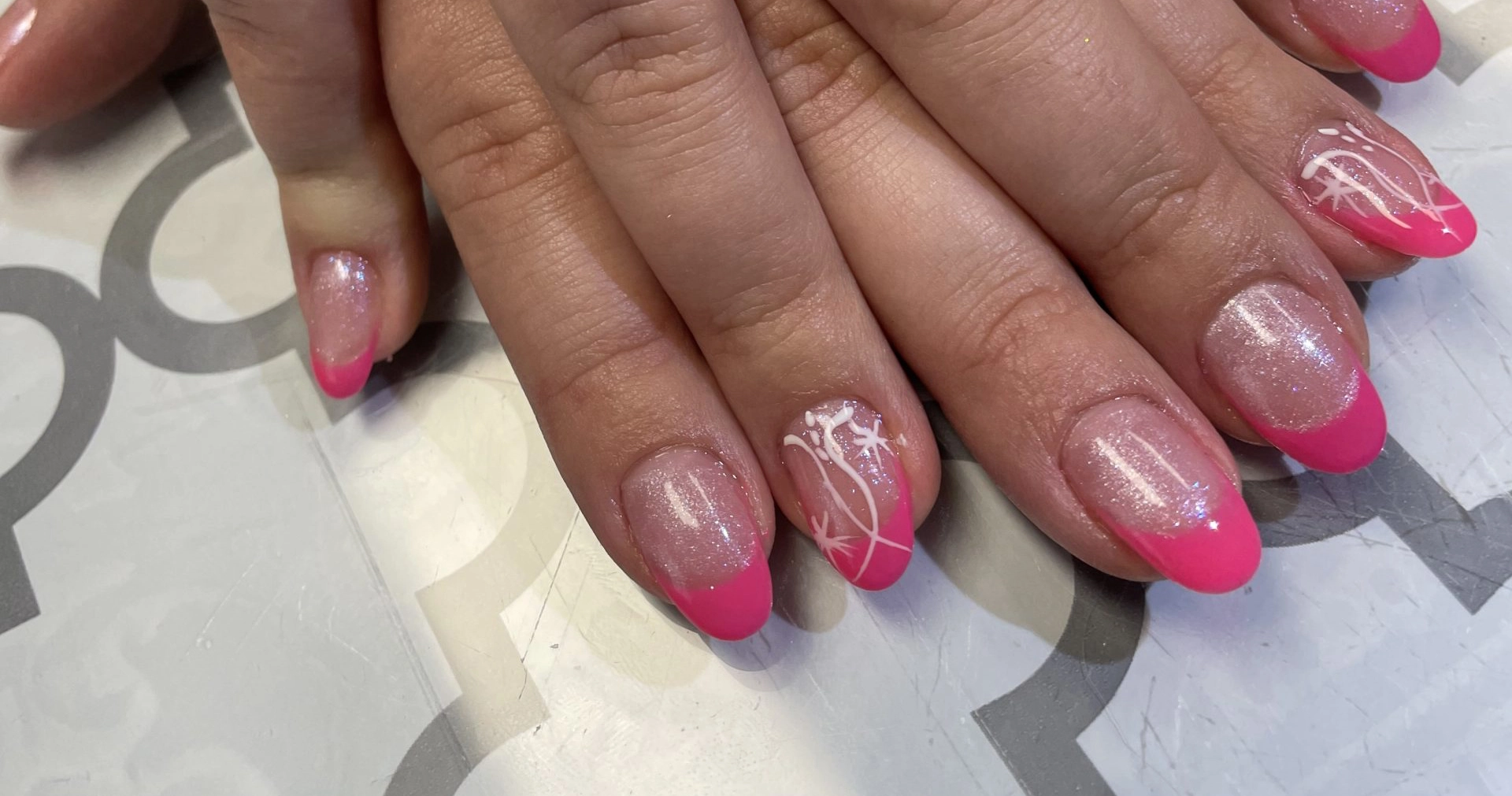 Closeup image of a woman's nails. The base of the nails is glittery, the tips are hot pink, and a couple nails have a white design.