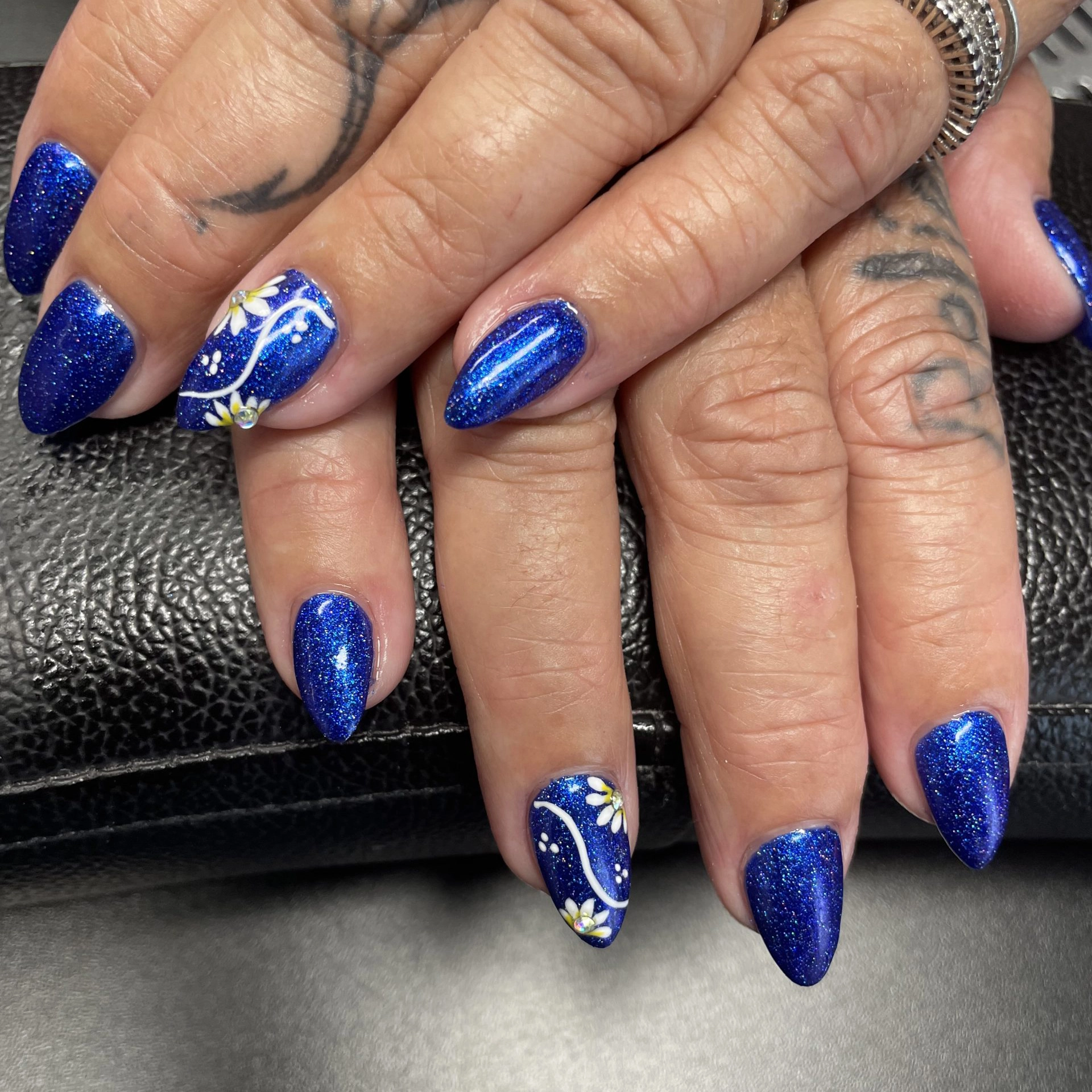 Closeup of a woman's dark blue glittery nails with a white flower design on two of the nails.